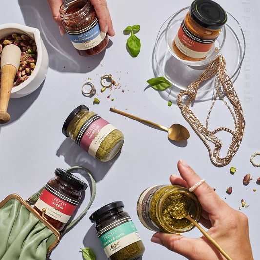 Five glass jars of Greenomic pesto varieties arranged on a light blue surface. The jars have black lids and labels with product names like "pesto calabrese" and "pesto arrabbiata". Surrounding the jars are various props including a mortar and pestle, jewellery, and a spoon.