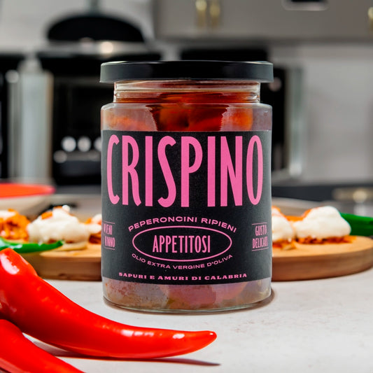 A glass jar of Crispino Appetitosi with a black label and pink text in a lifestyle setting. Within the image there are chilli's and peppers in a kitchen setting.
