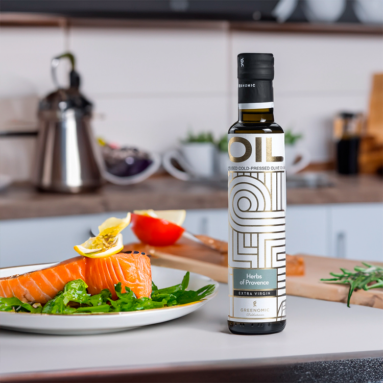A bottle of Greenomic Herbs of Provence Extra Virgin Olive Oil sits on a kitchen counter next to a plate of salmon and salad drizzeled in olive oil. The bottle is clear with a black cap and a white and gold label.
