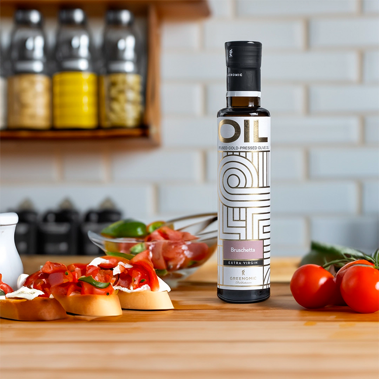 A glass bottle of Greenomic Bruschetta Extra Virgin Olive Oil on a wooden countertop. The bottle has a white label with gold accents and the words "Bruschetta" and "Extra Virgin" prominently displayed. A bowl of bruschetta with tomatoes and basil sits in the background, along with other ingredients like tomatoes and herbs.