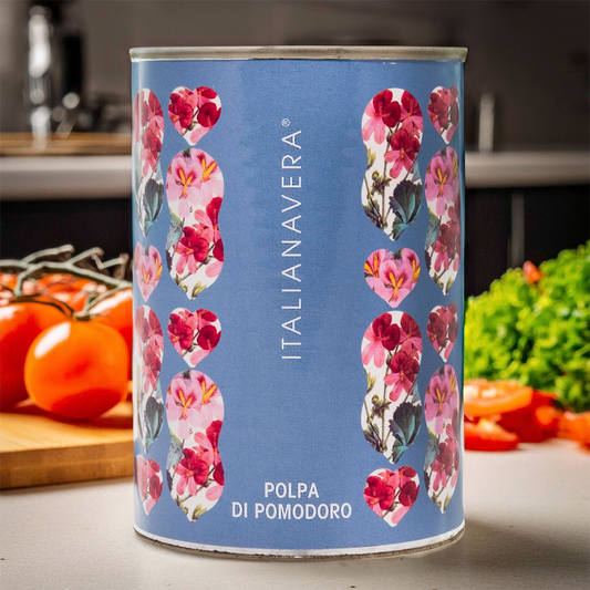 Itailanavera logo on a blue tin with pink botanical flowers. Image in a kitchen setting with tomatoes and herbs blurred in the background.