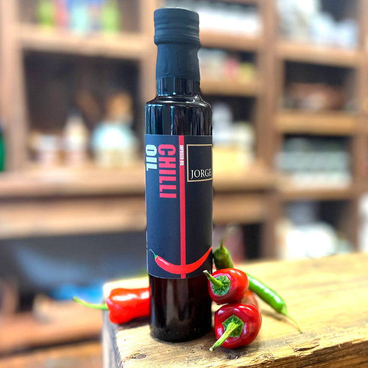 The glass chilli oil bottle in the Jorge's shop. In the image the chilli oil bottle is on a block of wood with red and green chillies resting on the bottle.