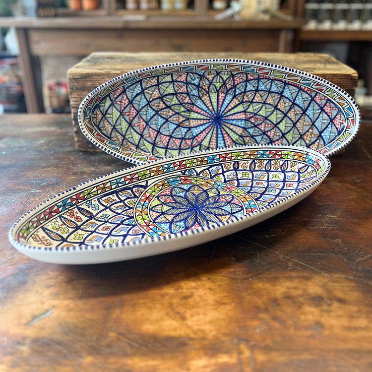 Two oval ceramic dishes displayed on a wooden table. The dishes feature colorful kaleidoscope patterns with intricate shapes and vibrant colors.