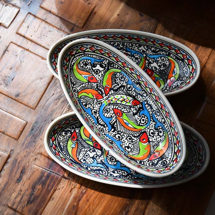Three oval ceramic dishes stacked on top of each other. Each dish features a colorful design of fish swimming among bubbles and waves. The fish are depicted in various shades of blue, green, orange, and red. The background of the dishes is white with intricate patterns. The dishes are resting on a wooden table.