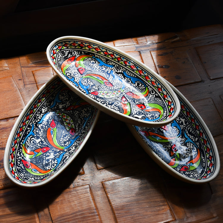 Three oval ceramic dishes stacked on top of each other. Each dish features a colorful design of fish swimming among bubbles and waves. The fish are depicted in various shades of blue, green, orange, and red. The background of the dishes is white with intricate patterns. The dishes are resting on a wooden table.