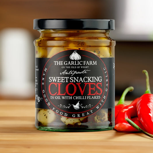 A glass jar with a black label with the garlic farm logo and the title of sweet snacking cloves in oil with chilli flakes in bold, placed on a wooden kitchen counter next to a selection of chillis