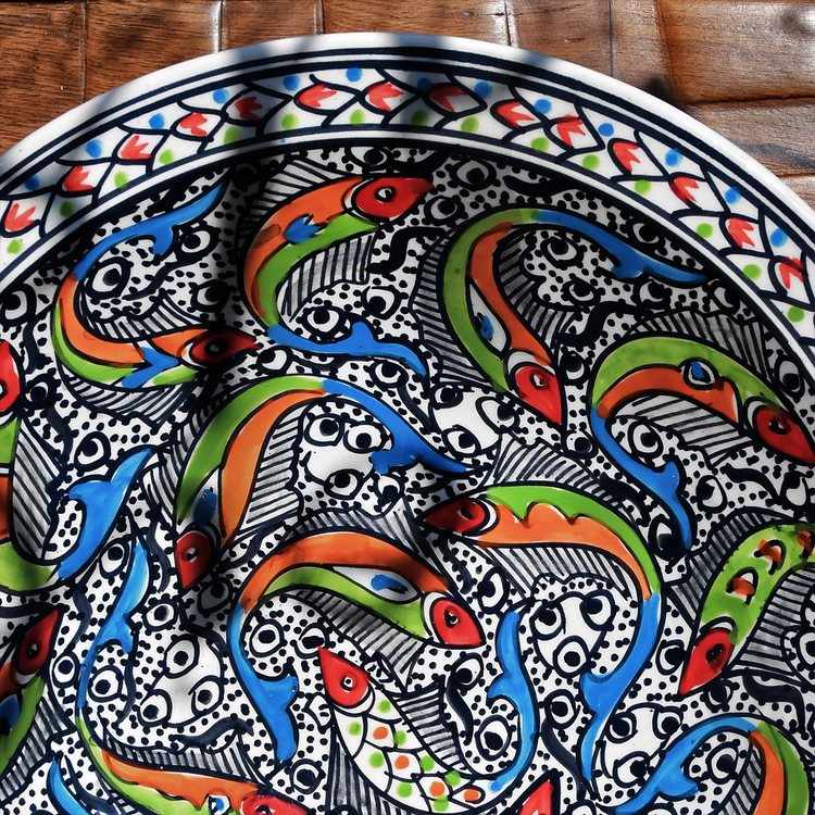 A circular ceramic plate with a vibrant, intricate design featuring multiple colorful fish swimming in a pattern against a white background. The fish have black outlines, orange and green scales, and black eyes. The plate has a border of geometric shapes in red, orange, and green.