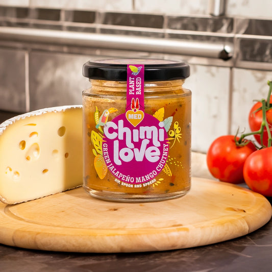 A jar of Chimi Love Medium Green Jalapeno & Mango Chutney jar on a cheeseboard next to cheese and tomatoes