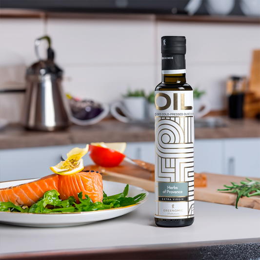 A bottle of Greenomic Herbs of Provence Extra Virgin Olive Oil sits on a kitchen counter next to a plate of salmon and salad drizzeled in olive oil. The bottle is clear with a black cap and a white and gold label.