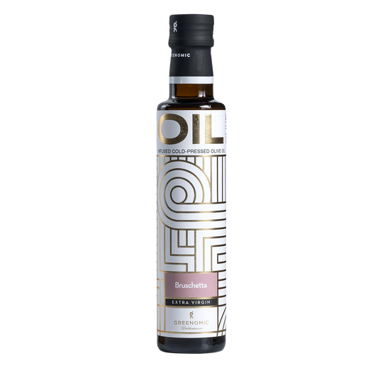 A glass bottle of Greenomic Bruschetta Extra Virgin Olive Oil on a wooden countertop. The bottle has a white label with gold accents and the words "Bruschetta" and "Extra Virgin" prominently displayed