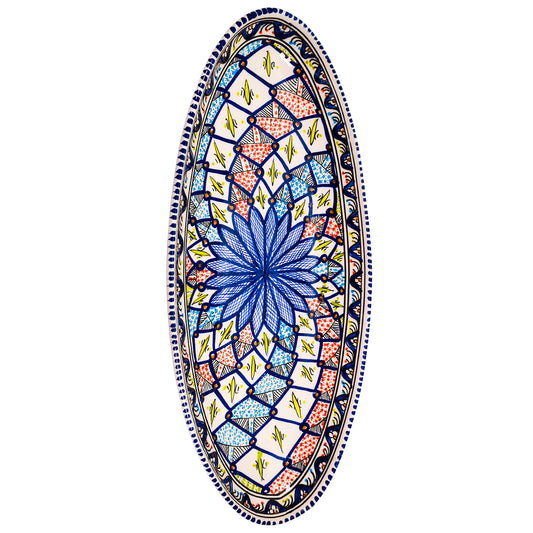 An oval ceramic dish with a colorful kaleidoscope pattern. The design features a central blue flower surrounded by intricate shapes in shades of red, pink, yellow, and blue. The border of the dish is decorated with a wavy line.
