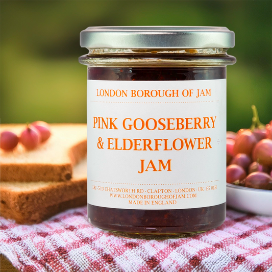 A clear jar with the London Borough of Jam logo and the title Raspberry & Hibiscus Flower Jam  on a white label with orange writing in a picnic setting 