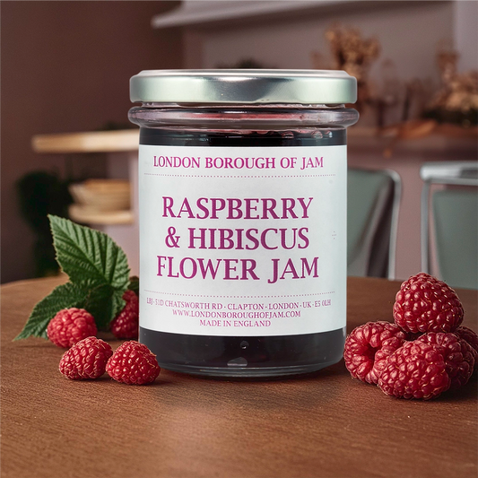 Glass jar of London Borough of Jam Raspberry & Hibiscus Flower Jam on a wooden table. The jar has a silver lid and is filled with a deep red jam. Fresh raspberries are scattered around the jar. The label includes the product name, ingredients, weight, and contact information.